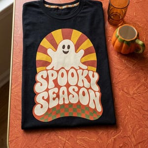 May include: A black t-shirt featuring a graphic print of a white ghost centered above the text "SPOOKY SEASON" in a bold, wavy font. The design includes a yellow and orange sunburst background and a green and orange checkered pattern at the base.