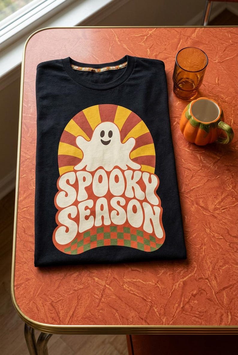 May include: A black t-shirt featuring a graphic print of a white ghost centered above the text "SPOOKY SEASON" in a bold, wavy font. The design includes a yellow and orange sunburst background and a green and orange checkered pattern at the base.