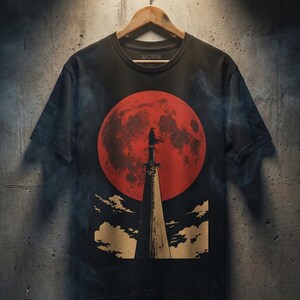 May include: A black short-sleeve t-shirt featuring a graphic print of a figure standing atop a tall, pointed structure against a large, textured red moon. The design includes stylized clouds at the base and the word WORN printed inside the neck collar.