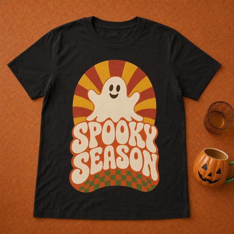 May include: A black short-sleeve t-shirt featuring a graphic print of a white ghost centered above the text "SPOOKY SEASON" in a bold, wavy font. The design includes a sunburst pattern in orange and yellow, with a green and orange checkerboard base.