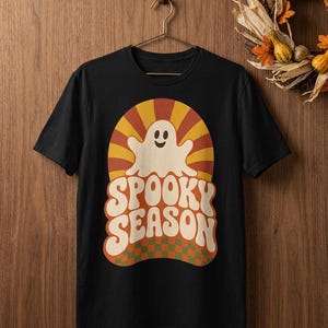 May include: A black short-sleeve t-shirt featuring a graphic print of a smiling white ghost centered within a sunburst pattern of orange and yellow rays. Below the ghost, the text SPOOKY SEASON appears in large cream letters over a green and orange checkerboard base.