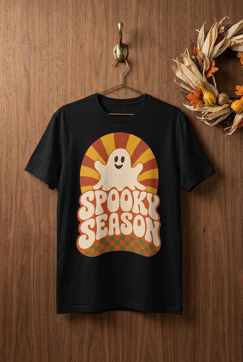 May include: A black short-sleeve t-shirt featuring a graphic print of a smiling white ghost centered within a sunburst pattern of orange and yellow rays. Below the ghost, the text SPOOKY SEASON appears in large cream letters over a green and orange checkerboard base.