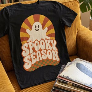 May include: A black short-sleeve t-shirt featuring a graphic print of a white ghost centered over a yellow and orange sunburst pattern. Below the ghost, the text SPOOKY SEASON is printed in a bold, wavy font above a green and orange checkered base.