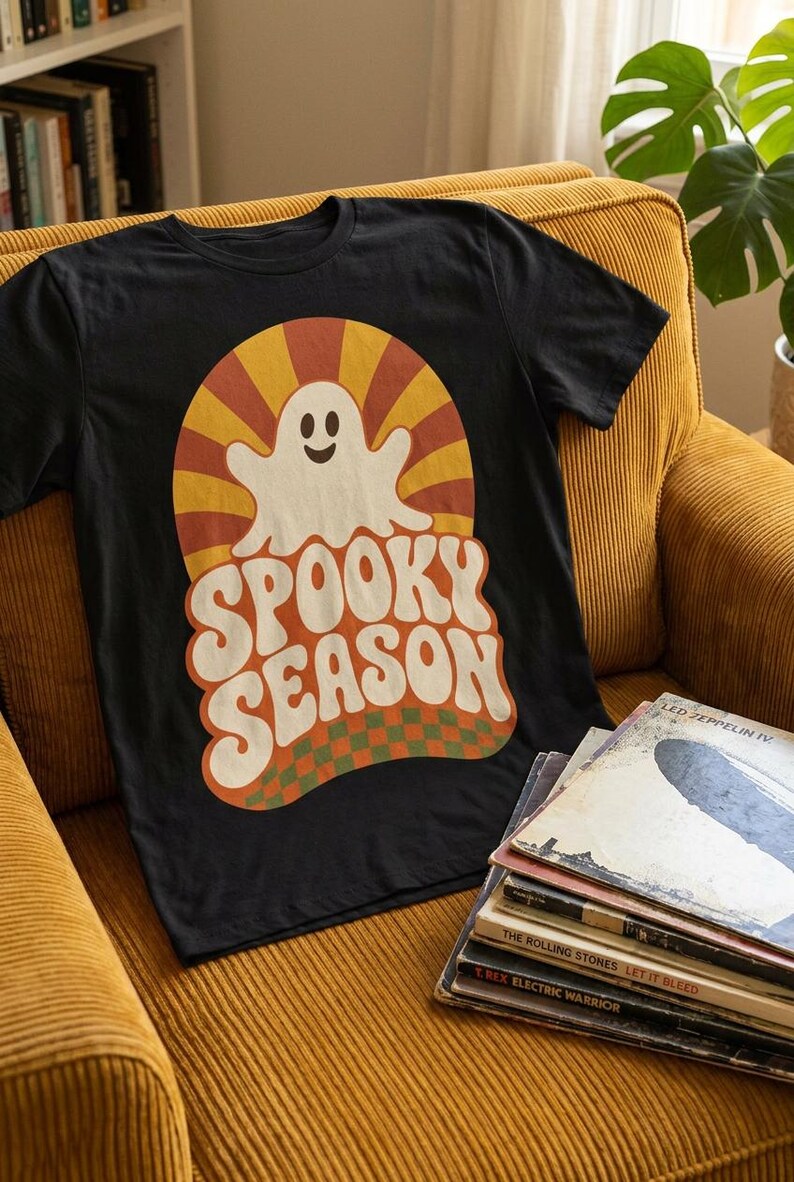 May include: A black short-sleeve t-shirt featuring a graphic print of a white ghost centered over a yellow and orange sunburst pattern. Below the ghost, the text SPOOKY SEASON is printed in a bold, wavy font above a green and orange checkered base.