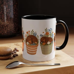 Puede incluir: Una taza de cerámica blanca con interior y asa negros, que presenta una ilustración repetida de perros salchicha marrones durmiendo dentro de varias macetas llenas de flores naranjas, azules y blancas y follaje verde.
