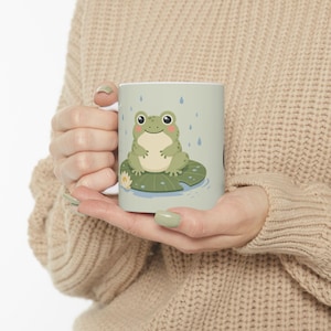 Puede incluir: Una taza de cerámica de color verde claro con una ilustración gráfica de una pequeña rana verde sentada sobre un nenúfar bajo la lluvia. El diseño incluye una flor de nenúfar blanca y gotas de lluvia azules sobre un fondo verde pálido.