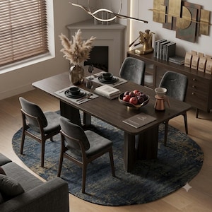 Op de afbeelding: Een donkerhouten eettafel met vier grijze gestoffeerde stoelen op een rond blauw en goudkleurig tapijt. De tafel is gedekt met zwarte borden, een schaal met granaatappels, een glazen vaas met droogbloemen en een koffiefilterhouder.