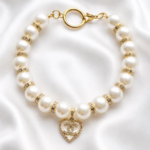 May include: A gold-toned bracelet featuring a series of white faux pearls separated by small, clear rhinestone-encrusted spacers. A heart-shaped pendant with a central interlocking CC logo and a rhinestone border hangs from the center of the chain.