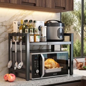 May include: A black metal two-tier kitchen storage rack holding a microwave with a roasted chicken inside, a rice cooker, glass spice jars, and bottles. Three silver metal ladles hang from the side hooks of the frame, and two red apples sit in front.