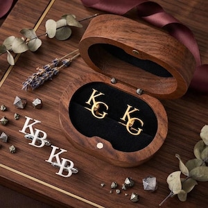 Op de afbeelding: Een paar goudkleurige manchetknopen met de initialen KG in een houten doosje, naast een paar zilverkleurige manchetknopen met de initialen KB, verspreide kleine metalen steentjes en gedroogde botanische takjes op een houten ondergrond.