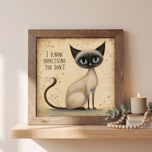 May include: A square framed wall art piece featuring a Siamese cat with large eyes and a long tail. The cream-colored background includes swirling line art, small stars, and the text "I KNOW SOMETHING YOU DON'T." The frame is made of dark brown wood.