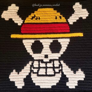 Pu&ograve; includere: Una coperta all'uncinetto nera con un disegno di teschio e ossa incrociate bianco e un cappello di paglia giallo con una fascia rossa. Il testo @knot_jo_mommas_crochet &egrave; visibile in alto. La coperta ha frange bianche, rosse e gialle sui lati.