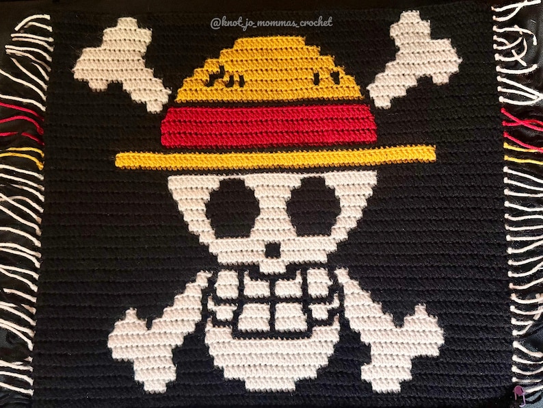 Pu&ograve; includere: Una coperta all'uncinetto nera con un disegno di teschio e ossa incrociate bianco e un cappello di paglia giallo con una fascia rossa. Il testo @knot_jo_mommas_crochet &egrave; visibile in alto. La coperta ha frange bianche, rosse e gialle sui lati.