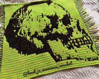 Può includere: Una coperta quadrata all'uncinetto con un design a teschio nero ad alto contrasto su uno sfondo verde neon vibrante. Il pezzo presenta un bordo sfrangiato con fili alternati neri e verde neon e include il testo @knotjo_mommas_crochet.