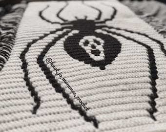 Può includere: Un tessuto rettangolare all'uncinetto con una grafica di ragno nero su sfondo bianco. L'addome del ragno mostra un motivo bianco simile a un teschio. Il testo @knot_jo_mommas_crochet è visibile a sinistra. I bordi presentano frange nere e bianche.