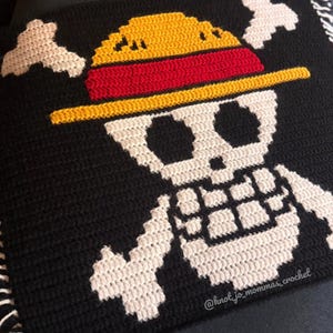 Pu&ograve; includere: Un quadrato all'uncinetto nero con un disegno bianco di teschio e ossa incrociate. Il teschio indossa un cappello di paglia giallo con una fascia rossa. Il testo @knot_jo_mommas_crochet &egrave; ricamato in bianco nell'angolo in basso a destra.