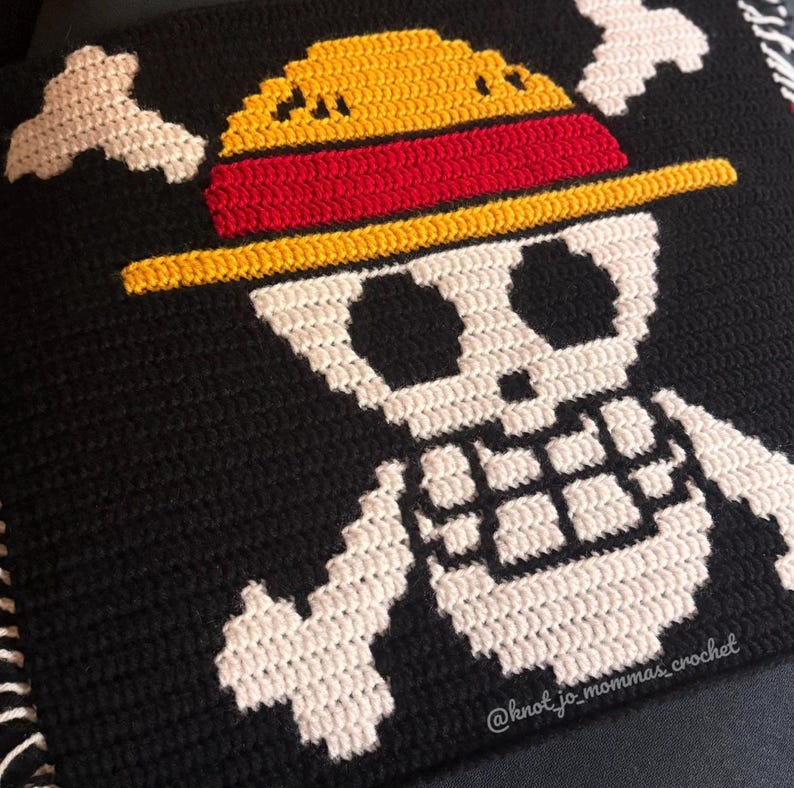 Pu&ograve; includere: Un quadrato all'uncinetto nero con un disegno bianco di teschio e ossa incrociate. Il teschio indossa un cappello di paglia giallo con una fascia rossa. Il testo @knot_jo_mommas_crochet &egrave; ricamato in bianco nell'angolo in basso a destra.