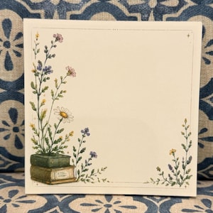 May include: A square notepad featuring a cream-colored background with a thin border. The illustration depicts two stacked green books with wildflowers growing from the top, accompanied by additional floral sprigs in the bottom right corner.