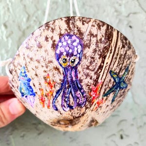 May include: A brown coconut shell bowl featuring a hand-painted purple octopus with white spots, a blue seashell, orange coral, and a blue starfish. The bowl has white string attached to the sides for hanging, showcasing a natural textured surface.