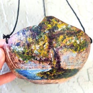 May include: A curved, bowl-shaped object made from a coconut shell, featuring a hand-painted landscape scene of a large tree with yellow and green foliage beside a blue body of water. The shell is suspended by thin black strings attached to the sides.