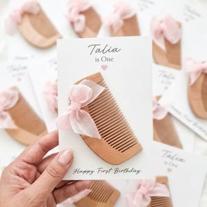 Könnte beinhalten: Ein hellbrauner Holzkamm, verziert mit einer transparenten rosa Schleife, befestigt auf einer weißen Karte mit dem aufgedruckten Text Talia is One und Happy First Birthday sowie einem kleinen rosa Herzsymbol über dem Namen.