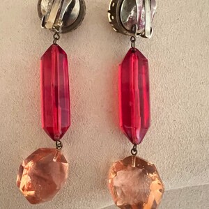 May include: A pair of dangle earrings with a silver tone clip-on closure. The earrings feature a red faceted teardrop-shaped bead and a clear faceted octagon-shaped bead.