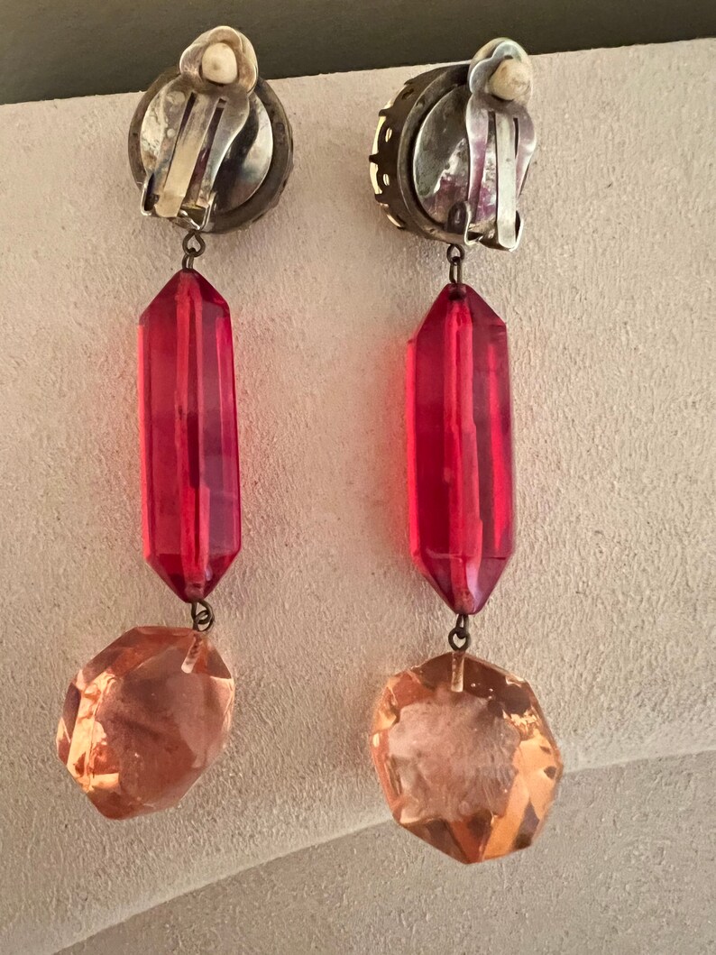 May include: A pair of dangle earrings with a silver tone clip-on closure. The earrings feature a red faceted teardrop-shaped bead and a clear faceted octagon-shaped bead.