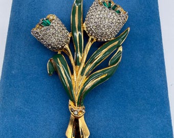 May include: A gold-toned brooch in the shape of a tulip bouquet. The tulip heads are encrusted with clear rhinestones and have green accents. The leaves are green with gold outlines, and the stems are gold. The brooch is set against a blue background.