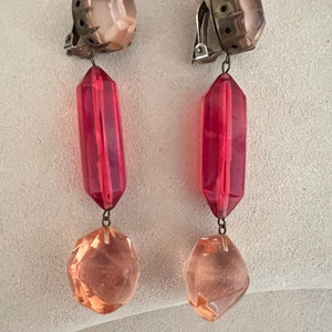 May include: A pair of clip-on earrings with a pink and red crystal design. The earrings feature a large pink crystal at the top, followed by a long red crystal, and a smaller pink crystal at the bottom.