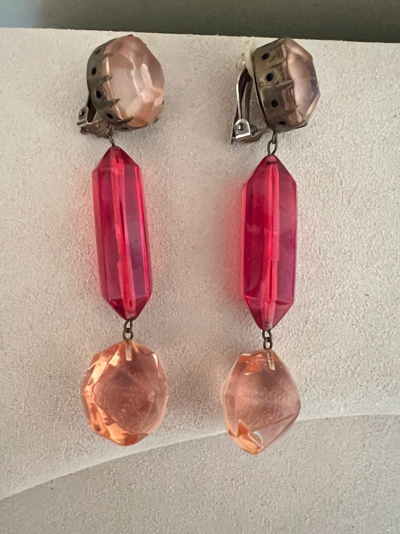 May include: A pair of clip-on earrings with a pink and red crystal design. The earrings feature a large pink crystal at the top, followed by a long red crystal, and a smaller pink crystal at the bottom.