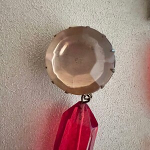 May include: A red crystal pendant with a clear glass bead at the top. The pendant is attached to a silver-toned metal loop.