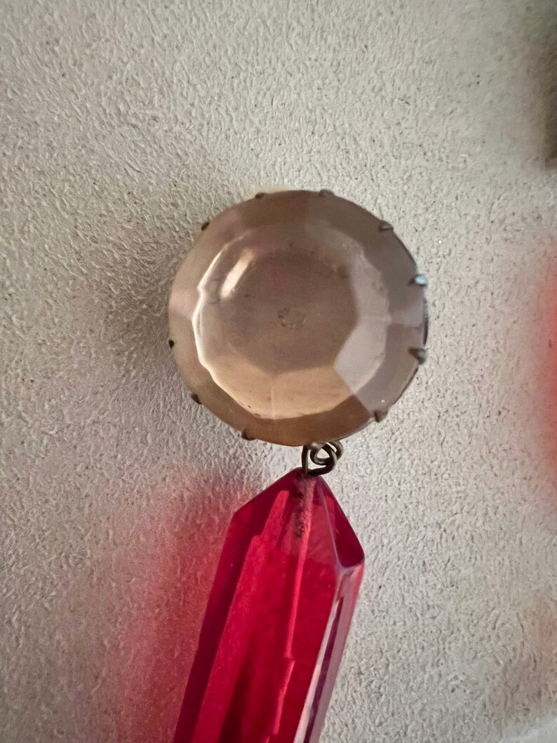 May include: A red crystal pendant with a clear glass bead at the top. The pendant is attached to a silver-toned metal loop.