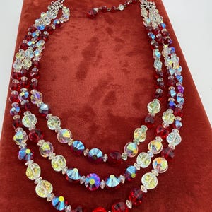 May include: A three-strand necklace with red and iridescent beads. The necklace features a variety of bead shapes, including round and faceted, with a silver-tone clasp. The necklace is displayed on a red velvet surface.