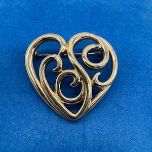 May include: A gold-toned heart-shaped brooch with an openwork design. The brooch features swirling, stylized details within the heart's outline. The pin is set against a blue background.