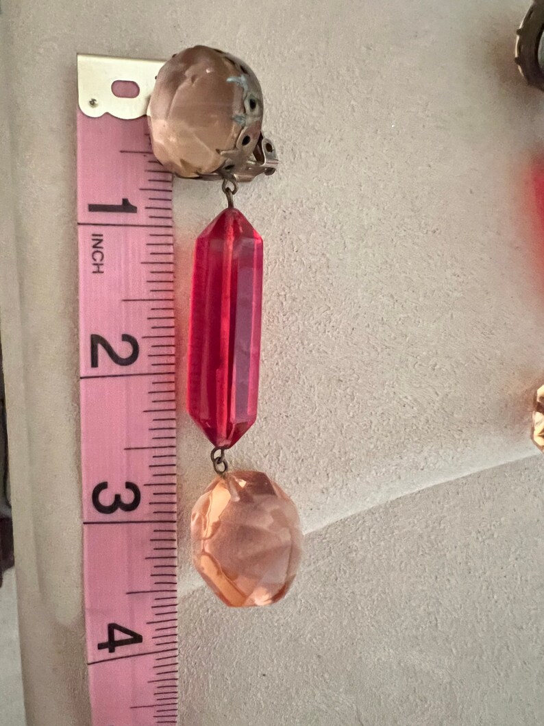 May include: A pair of dangle earrings with a red faceted crystal and a peach-colored faceted crystal. The earrings are approximately 3 inches long.