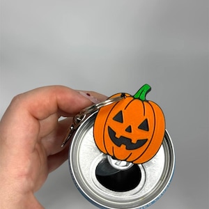 Op de afbeelding: Een oranje pompoenvormige blikopener met een groene steel en een zwart jack-o'-lantern gezicht. De opener is bevestigd aan een zilveren metalen sleutelhanger en is geplaatst over de opening van een blauw aluminium drankblikje.