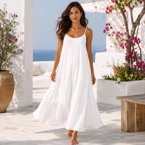 May include: A floor-length white maxi dress featuring a scoop neckline and thin spaghetti straps. The garment has a loose, flowing silhouette with a gathered bodice that extends into a full, pleated skirt that reaches the ankles.