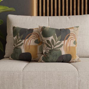 May include: Two square throw pillows featuring an abstract print with olive green, mustard yellow, and dark charcoal shapes. The design includes a continuous line drawing of a face, botanical leaf motifs, and arched lines on a light beige background.