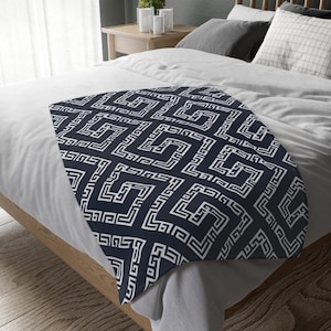 Puede incluir: Una manta de color azul marino oscuro con un patrón geométrico griego repetitivo en blanco. La manta está extendida sobre una cama con ropa de cama blanca y gris, mostrando el intrincado diseño lineal sobre la superficie de tela suave.