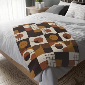 Puede incluir: Una manta con un patrón geométrico de cuadrados en tonos marrón oscuro, crema, amarillo mostaza y terracota. El diseño incluye formas circulares abstractas y finas líneas de cuadrícula negras sobre los bloques de color sólido.