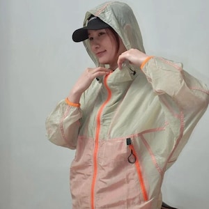 May include: A lightweight, translucent hooded windbreaker jacket featuring a color-blocked design with a pale green upper section and a soft peach lower section. The jacket includes a bright orange front zipper, orange trim, and a black zipper pull.