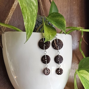May include: A pair of drop earrings featuring three circular, dark brown, textured discs of varying sizes connected by silver-toned metal jump rings. Each disc has two small holes in the center, resembling buttons, and hangs vertically from a silver-toned hook.