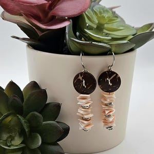 May include: A pair of dangle earrings featuring dark brown circular wooden buttons at the top, connected to silver-toned hooks. Below each button hangs a vertical stack of small, pale pink shell discs interspersed with tiny silver-toned metal beads.