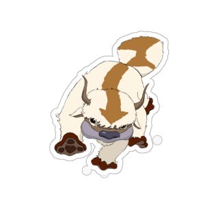 May include: A die-cut sticker featuring a cartoon creature with cream-colored fur, brown markings on its back and tail, curved horns, and a dark nose. The creature is depicted in a forward-facing pose with visible paws and a fluffy tail.