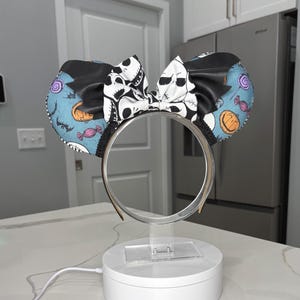 May include: A headband featuring two circular ears with a light blue background patterned with orange pumpkins, purple candies, and black bats. A central black and white bow displays a repeating skull face print, and the headband has a thin frame.
