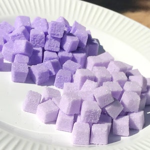 May include: Two piles of small, lavender-colored sugar cubes on a white plate.