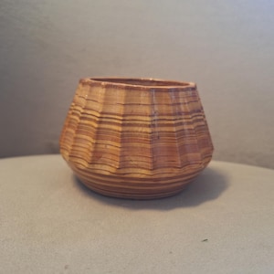 Peut inclure: Un petit bol rond avec une surface extérieure cannelée et plissée. L'objet présente un motif de grain de bois distinct avec des bandes horizontales alternées de couleurs beige clair, brun moyen et brun foncé sur tout l'extérieur.