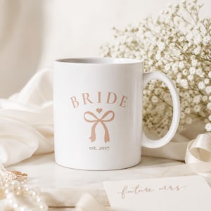 Könnte beinhalten: Eine weiße Keramiktasse mit dem Text BRIDE in geschwungener Schrift über einer hellrosa Schleifengrafik und dem Text est. 2027. Die Tasse hat einen Standardhenkel und eine glatte Oberfläche mit einem klaren Design auf der Vorderseite.