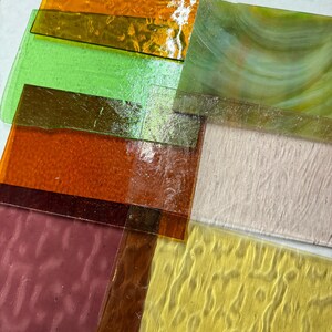 May include: Assortment of textured glass sheets in various colors, including green, orange, yellow, and brown. The sheets are stacked on top of each other, with the top sheet being a light green with a swirl pattern.
