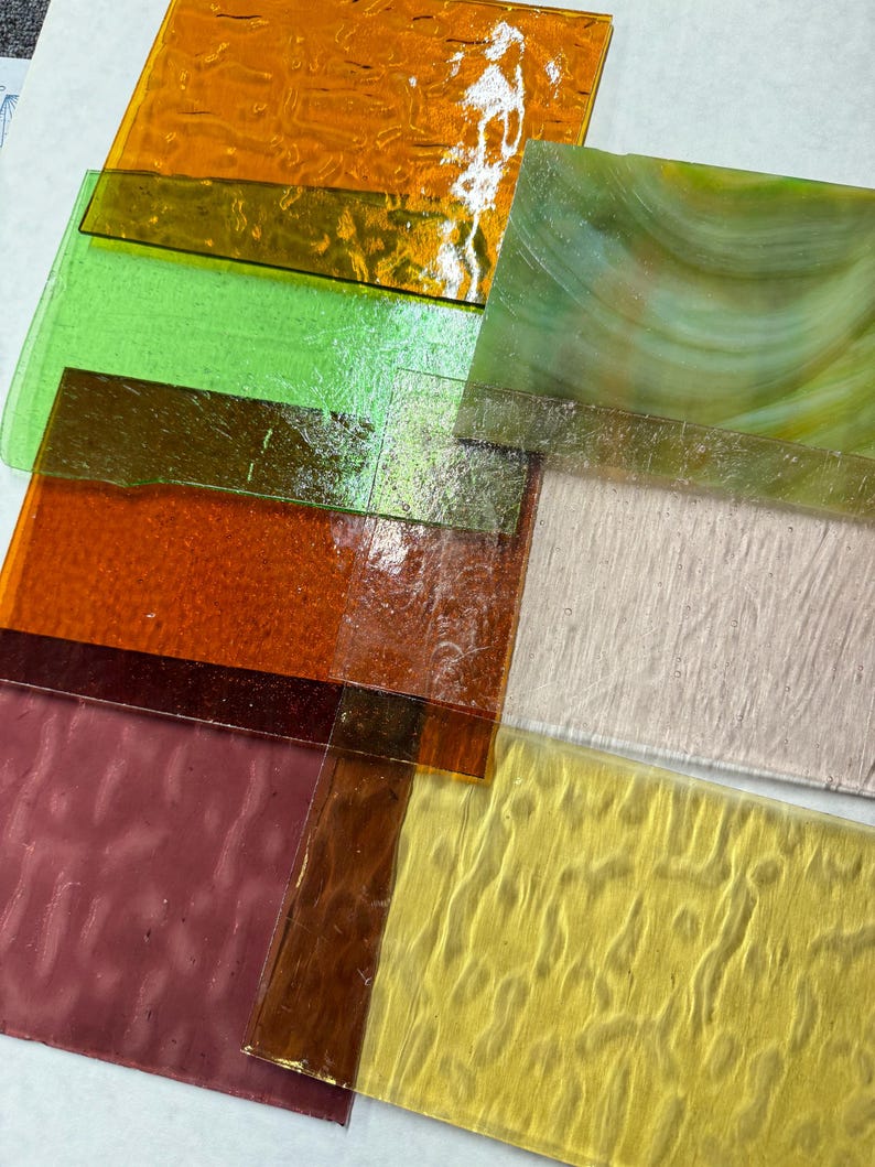 May include: Assortment of textured glass sheets in various colors, including green, orange, yellow, and brown. The sheets are stacked on top of each other, with the top sheet being a light green with a swirl pattern.